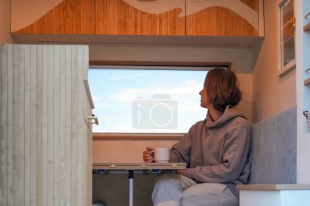 A young man enjoys a peaceful moment with a warm drink inside his self-converted van, showcasing a simplistic and intimate lifestyle against the backdrop of a fading evening light.