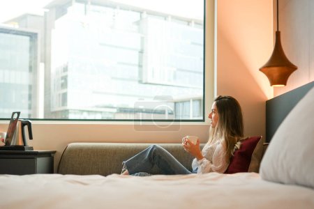 Une jeune femme est assise confortablement près d'une fenêtre dans une chambre d'hôtel, tenant une tasse avec une toile de fond urbaine sereine, suggérant un moment de détente ou de contemplation.