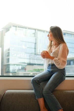 Eine junge Frau sitzt gelassen am Fenster eines hellen Hotelzimmers, blickt nach draußen und verkörpert ein Gefühl von Ruhe und Komfort, während sie die Aussicht genießt..