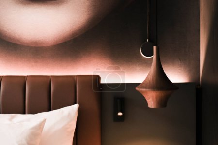 Dieses Bild zeigt einen Teil eines modernen Hotelzimmers mit einem plüschigen Kopfteil, einer eleganten Pendelleuchte und einem künstlerischen Wanddekor, das einen Hauch von Raffinesse verleiht..