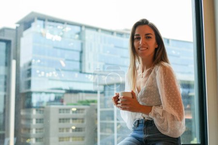 Une jeune femme invitée dans une chambre d'hôtel assise sur un rebord de fenêtre, tenant une tasse de café.