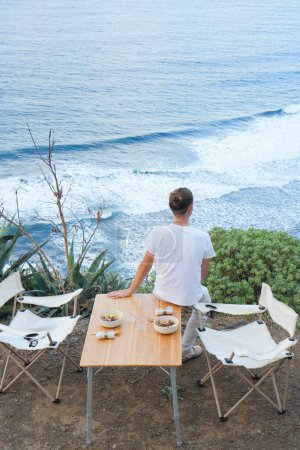 A man in solitude, seated at an outdoor dining setup on a cliff, gazes at the ocean during dusk, surrounded by natures beauty