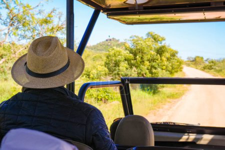 Un turista con sombrero de sol está sentado en un vehículo 4x4, explorando los paisajes naturales del Parque Nacional Kruger. El cielo despejado y el camino de tierra indican un viaje aventurero por delante, en busca de vida silvestre en una de las reservas de caza más grandes de África.
