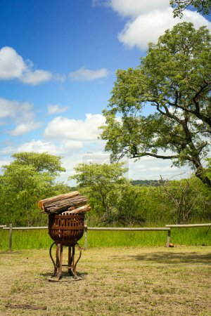 Ein rustikaler Holzwagen steht in einer ruhigen Landschaft, ein großer Baum spendet an einem sonnigen Tag Schatten. Die Szenerie ist friedlich und unterstreicht den Charme des ländlichen Lebens mit einem klaren blauen Himmel im Hintergrund und einem sattgrünen Baum, der das c