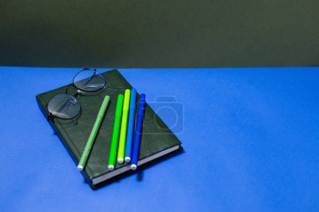 Photo for Dark green notebook with glasses and multi-colored felt-tip pens on a blue background - Royalty Free Image