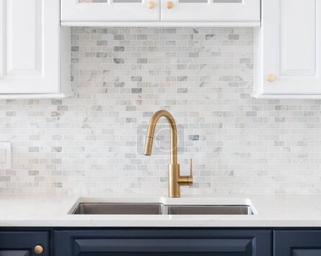 Photo for A kitchen sink detail shot in a white and blue kitchen with a gold faucet, marble countertop, and small marble subway tile backsplash. - Royalty Free Image