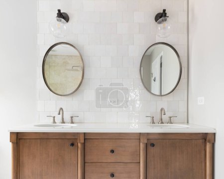 Foto de Un acogedor cuarto de baño con una vanidad de madera natural, salpicaduras de azulejos y luces montadas sobre espejos circulares. - Imagen libre de derechos