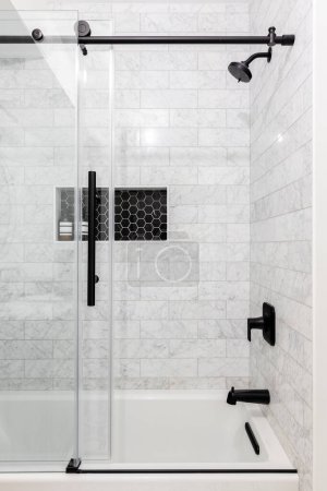 A remodeled glass shower with marble subway tiles, a shelf with black hexagon tiles, and a sliding glass door with black hardware.