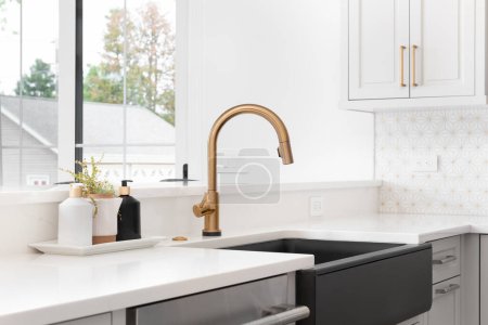 Téléchargez les photos : A beautiful sink in a remodeled modern farmhouse kitchen with a gold faucet, black farmhouse sink, white granite, and a tiled backsplash. No labels. - en image libre de droit