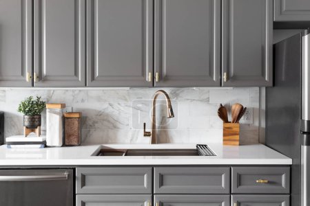 A kitchen sink detail shot with grey cabinets, a marble subway tile backsplash, and gold hardware and faucet.