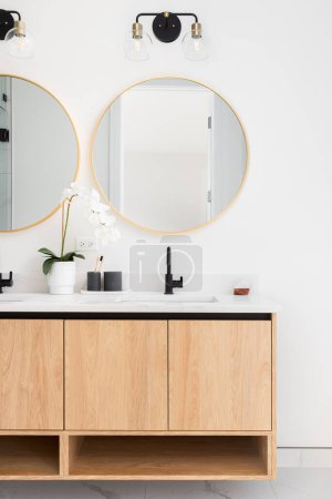 Foto de A beautiful bathroom with a floating wood cabinet, marble countertop, and gold circular mirror. - Imagen libre de derechos