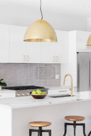 Photo for A kitchen detail with white cabinets, gold faucet and light hanging over the island with bar stools, and a tiled backsplash. - Royalty Free Image