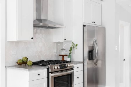 A kitchen detail with white cabinets, stainless steel appliances, tan hexagon tile backsplash, and decorations on the grey marble countertop.