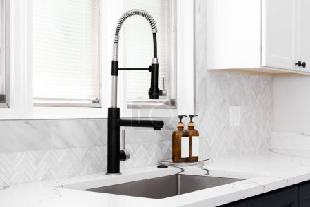 Photo for A kitchen sink detail shot with blue and white cabinets, marble herringbone tile backsplash, and a black faucet in front of a window. - Royalty Free Image