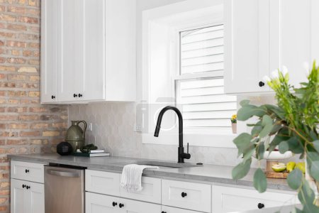 Photo for A beautiful kitchen sink detail with white cabinets, marble countertops, and a hexagon tiled backsplash. A blurred plant in the foreground. - Royalty Free Image