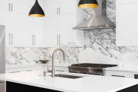 Photo for A kitchen detail with white cabinets, modern light fixtures hanging over a black island, and a marble countertop and backsplash. - Royalty Free Image