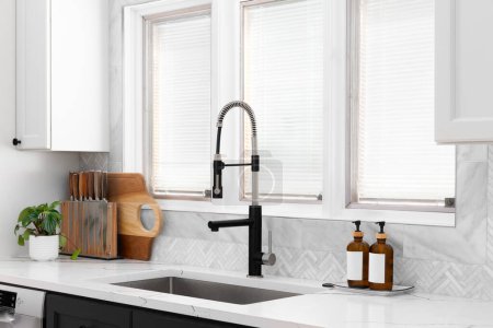 Photo for A kitchen sink detail with a herringbone tile backsplash, black and stainless steel faucet, white and black cabinets, and decor on the marble counter. - Royalty Free Image