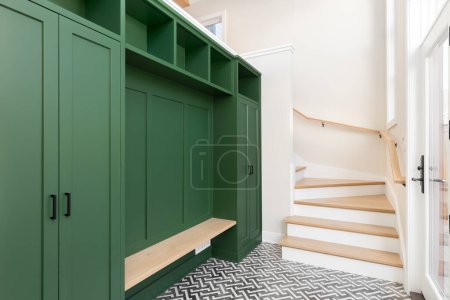 Un gran vestíbulo con una unidad de almacenamiento verde vibrante, banco de roble blanco y escaleras, y un suelo de baldosas en blanco y negro.