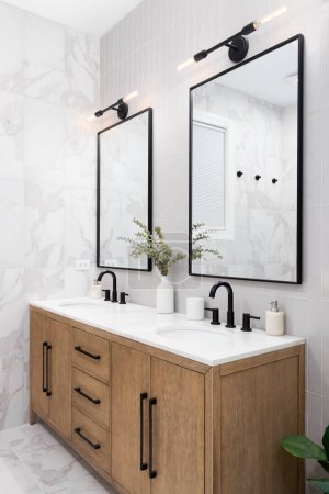 Photo for A bathroom with marble and stacked vertical subway tiles, a white oak vanity cabinet, black framed square mirrors and faucets, and marble countertop. - Royalty Free Image