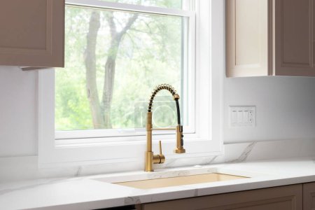 Photo for A gold kitchen faucet detail with a gold sink, brown cabinets, and a white marble countertop. - Royalty Free Image