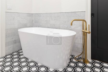Photo for A freestanding tub with a gold faucet, black and white pattern tile flooring, marble subway tile backsplash, and a black sliding door. - Royalty Free Image