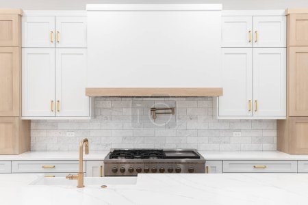 Photo for A kitchen detail with gold faucet and hardware, a marble subway tile backsplash, a large stove hood, and white and white oak cabinets. - Royalty Free Image
