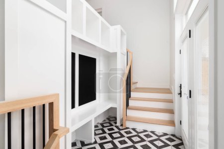 A large foyer with a white storage unit, white bench, white oak stairs and railing, and a black and white pattern tile flooring.