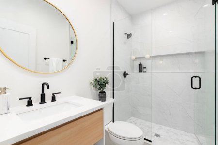 Ein modernes Badezimmer mit einem weißen Eichenschrank mit einer Arbeitsplatte aus weißem Marmor, einem runden Spiegel aus Gold und einer Dusche mit sechseckigen und rechteckigen Fliesen aus Marmor.