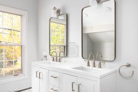 Foto de Un detalle del cuarto de baño con un gabinete blanco, grifos y espejos de bronce, y árboles coloridos por las ventanas. - Imagen libre de derechos