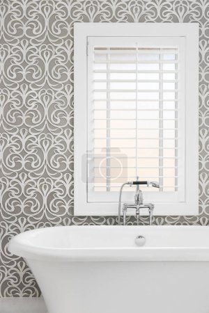 Téléchargez les photos : Un détail de baignoire autoportante avec un mur gris et blanc motif tuile, robinet chromé poli, et volets blancs sur la fenêtre. - en image libre de droit