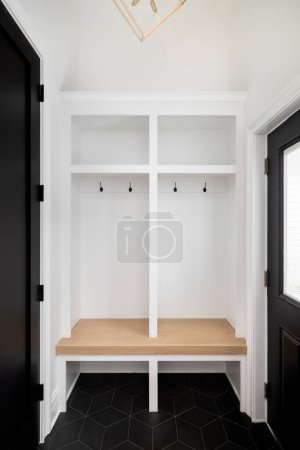 Une armoire d'entrée avec étagères blanches et banc en bois clair, carrelage noir, portes noires et un lustre en or suspendu au-dessus.