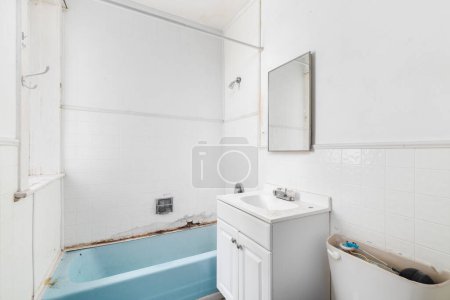 A bathroom with water damage and falling apart with white square tiles, a white bathroom cabinet, and a dated blue bathtub ready to be renovated.