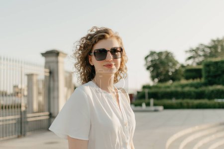 Foto de Una mujer con el pelo rizado en gafas de sol y un vestido blanco posando en el parque al atardecer. - Imagen libre de derechos