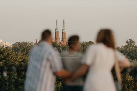 Foto de A photo from the back of father, mother, and son who are out of focus and staring at the old European church. The family of tourists is enjoying the town sight in the evening. Tourists at sunset. - Imagen libre de derechos
