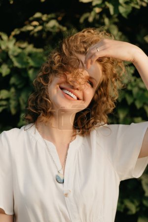 Foto de A smiling woman with curly hair is posing in the park in the evening. A close portrait of a lady in a white dress with green leaves in the background. - Imagen libre de derechos