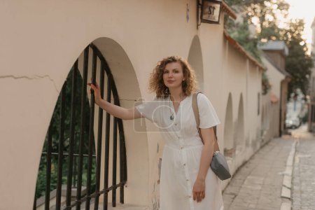 Téléchargez les photos : A woman with curly hair is holding on to a bar of a metal lattice in an old European town. A lady in a white dress in the evening. - en image libre de droit