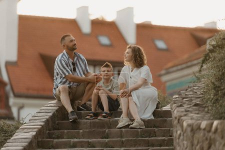 Téléchargez les photos : Une famille est assise sur les escaliers entre les toits d'une vieille ville européenne. Un père, une mère et un fils heureux s'amusent le soir. - en image libre de droit