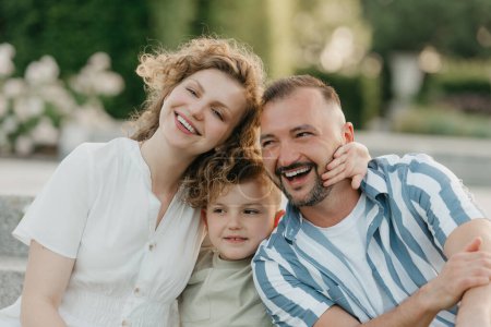 Téléchargez les photos : Father, mother, and son are sitting on the steps in the garden of an old European town. A close photo of a laughing family in the park in summer at sunset. - en image libre de droit
