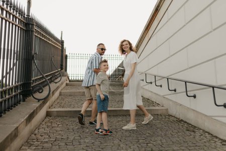 Photo for A photo from the back of a family is looking back while climbing the stairs of the palace in an old European town. A happy father, mother, and son are holding hands in the evening. - Royalty Free Image