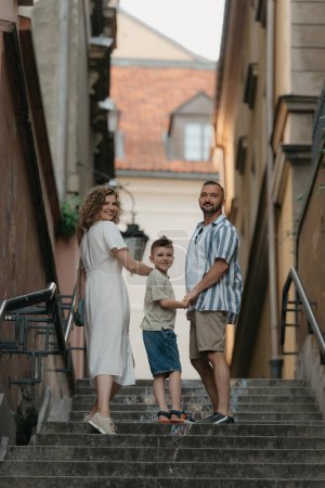 Téléchargez les photos : Une photo pleine longueur du dos d'une famille qui se retourne en montant des escaliers dans une vieille ville européenne. Un père, une mère et un fils heureux se tiennent la main et s'amusent le soir. - en image libre de droit