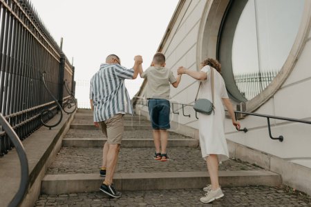 Téléchargez les photos : A photo from the back of a family is climbing the stairs of the palace in an old European town. A happy father, mother, and son are holding hands in the evening. - en image libre de droit