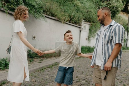 Téléchargez les photos : Father, mother, and smiling son are holding hands on the green cobbled street of an old European town. Happy family in the evening. Tourists at sunset. - en image libre de droit