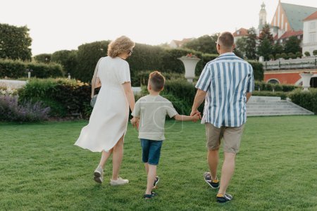 Téléchargez les photos : A photo from the back of a happy family is strolling the garden of the palace in an old European town. A smiling father, mother, and son are holding hands and having fun at sunset - en image libre de droit