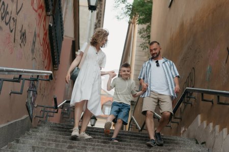 Foto de A full-length photo of a family which is going down the stairs in an old European town. A happy father, mother, and son are holding hands and having fun in the evening. - Imagen libre de derechos
