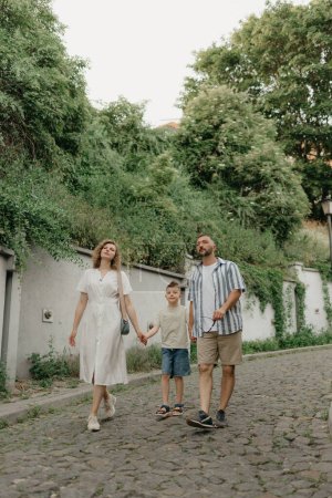 Photo for Father, mother and son are holding hands on the green cobbled street of an old European town. Happy family in the evening. Tourists at sunset. - Royalty Free Image