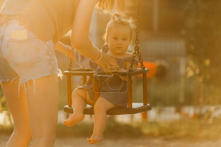 Téléchargez les photos : Bébé fille tout-petit sur une balançoire sur la chaude soirée d'été. Mère balance sa jeune fille sur une aire de jeux ensoleillée. - en image libre de droit