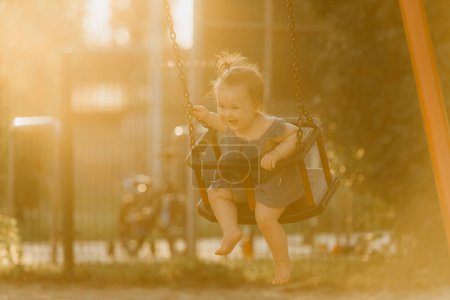 Téléchargez les photos : Bébé fille tout-petit sur une balançoire sur la chaude soirée d'été. Mère balance sa jeune fille sur une aire de jeux ensoleillée. - en image libre de droit