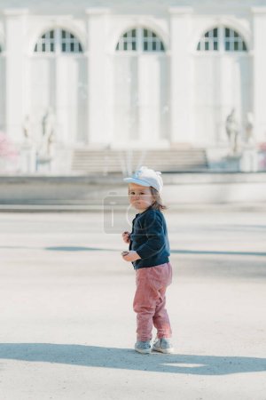 Photo for A female toddler in a cap and velvet overall with her back turns in the yard of the European palace. A baby girl walks to the fountain. - Royalty Free Image