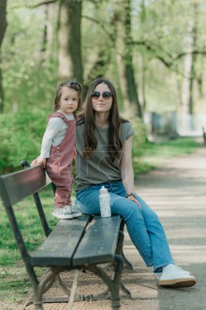Photo for A mother rests close to her little daughter on the bench in the park. A female toddler in velvet overall is having fun with her mom in the garden. - Royalty Free Image