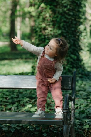 Téléchargez les photos : Un tout-petit femelle dans un ensemble de velours tend un cookie sur le banc de jardin. Une petite fille s'amuse dans le parc. - en image libre de droit